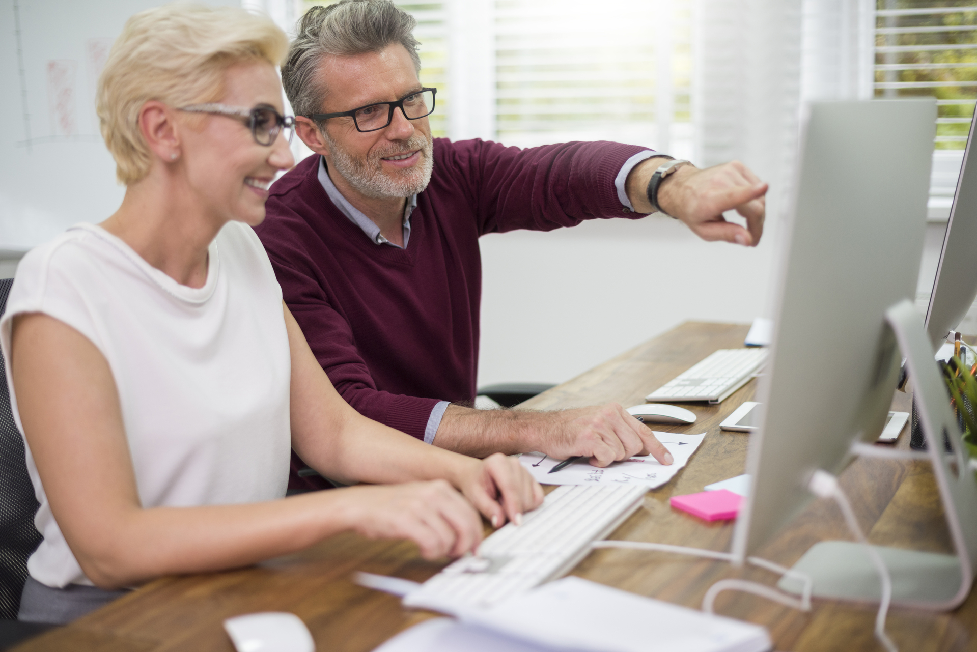 Deux seniors assis devant des ordinateurs, pointant du doigt l’écran et apprenant ensemble, illustrant le service formation numérique.